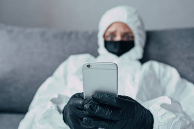 Virüs koruyucu kıyafetli, tıbbi maskeli ve siyah eldivenli bir virologun elinde bir telefon. İnternette kendinizi koronavirüsten nasıl koruyacağınızla ilgili bilgi arıyor.