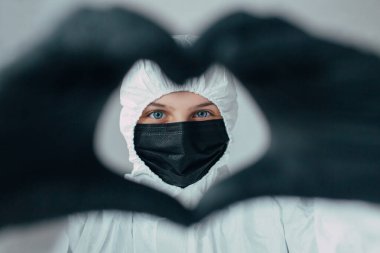 Beyaz koruyucu tıbbi maskeli bir doktor siyah eldivenli bir kalbi kameraya gösteriyor, koronavirüs salgınının sonuçlarını düşünüyor, insanları birleştiriyor ve birbirlerini seviyorlar.