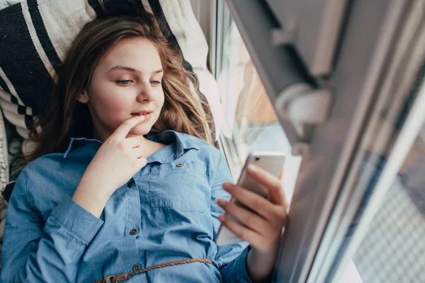 身穿蓝色衣服的少女躺在窗台上 看着电话 用手指捂着嘴笑着 — 图库照片