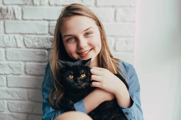 一个蓝眼睛的微笑的女孩抱着一只黑猫在白砖墙的后面 宠物和主人之间的友好关系 长有浅棕色长发的青少年坐在靠窗的地方 拥抱着她 — 图库照片