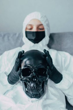 Viral koruyucu giysi ve tıbbi maske takmış bir hemşire elinde siyah bir kafatası tutuyor ve koronavirüs salgınının sonuçlarını endişeyle düşünüyor. Virüs yüzünden izole edilmiş..