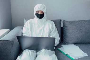 Viral koruyucu giysi ve siyah maske giyen bir doktor bir dizüstü bilgisayarda çalışıyor, internette koronavirüs salgını hakkında bilgi arıyor. Sokağa çıkma yasağı ve kapalı sınırlarda karantinaya alındı ve izole edildi