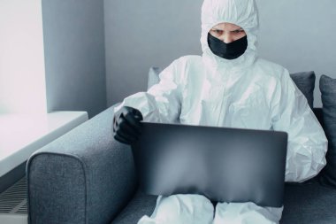 Yüz maskesi ve viral koruyucu giysili sıhhiye bir dizüstü bilgisayar kullanıyor ve Coronavirus salgınıyla ilgili haberleri araştırıyor. Sokağa çıkma yasağı zamanında karantinaya alınmış ve kendi kendini izole etmiş..