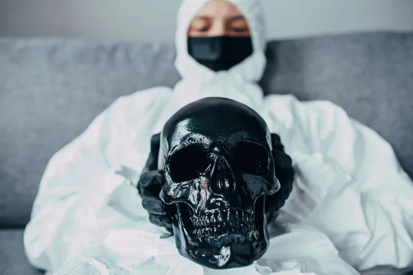 black skull holding by physician in face medical mask and viral protective suit. Medic look on it and think about consequences and mortality of coronavirus epidemic. Danger of death due the pandemic.