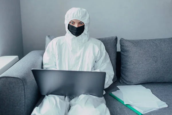 A physician in a viral protective suit and black mask works on a laptop, online searching information about  coronavirus pandemic. Quarantined and isolated at home in curfews time and closed border