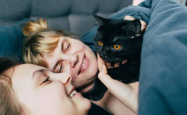mother and daughter sleeps together in a bed with a black cat. Family spending time together. Close up portrait of smiling woman holding cat on hands while laying on blue bed with her child at home clipart