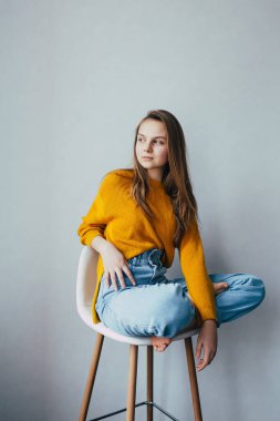 Genç kız beyaz, modern, beyaz sandalyeye oturmuş iki bacağını kaldırmış bir şekilde yan tarafa bakıyor. Evin içinde sarı kazaklı ve kot pantolonlu güzel bir kız. Moda kıyafetleri. 