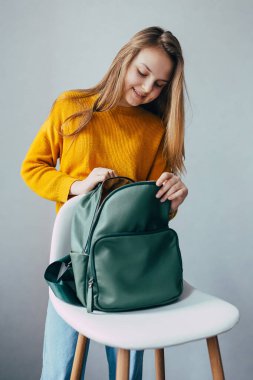 Genç kız, beyaz, modern bir sandalyede duran deri sırt çantalarının içine bakıyor. Sarı kazaklı ve kot pantolonlu güzel kız kal ve yeşil sırt çantasına bak. Moda kıyafetleri