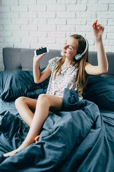 아름다운 소녀가 침대에서 현대식 헤드폰을 커다란 전화기를 음악을 활동적 — 스톡 사진