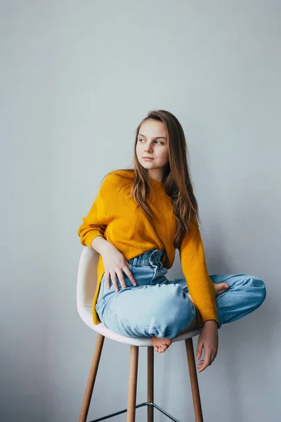 teenage girl looks at side sitting on white modern white chair with two leg raise up. Beautiful girl in yellow sweater and blue jeans in home interior. Trendy casual outfit.