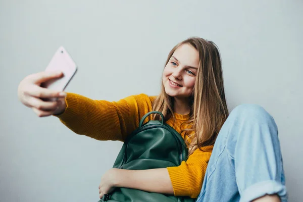 少女拿着手机自拍 对着相机笑 穿着黄色毛衣和蓝色牛仔裤的漂亮女孩拿着手机和绿色背包 时髦休闲装 — 图库照片