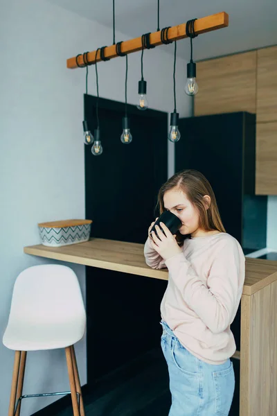美国少女在家里厨房里用黑杯喝咖啡 穿着粉色衬衫和蓝色牛仔裤的女人 现代室内设计及灯泡黑色金属丝吊灯 — 图库照片