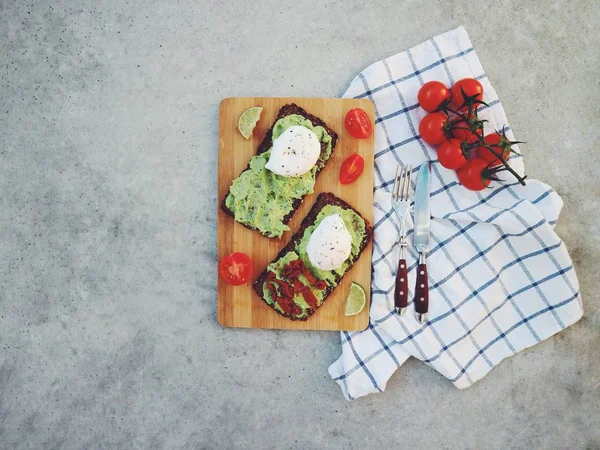 Top view of food composition of sandwiches with mashed avocado, poached eggs, lime slices and cherry tomatoes served with napkind and cutlery