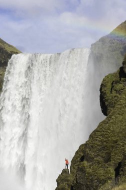 Kırmızı ceketli genç adamı Skogafoss Şelalenin arkasında, İzlanda bir uçurumun üzerinde duruyor.