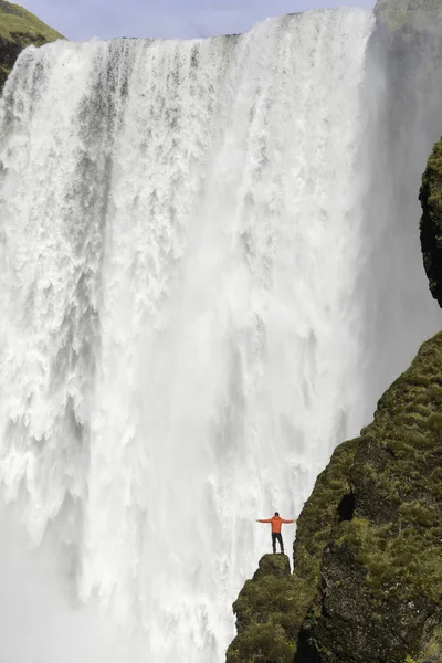 Junger Mann in roter Jacke steht auf einer Klippe mit skogafoss Wasserfall im Hintergrund, Island. — Stockfoto