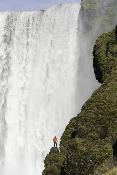 Junger Mann in roter Jacke steht auf einer Klippe mit skogafoss Wasserfall im Hintergrund, Island. — Stockfoto