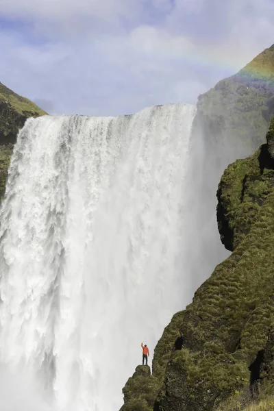 Junger Mann in roter Jacke steht auf einer Klippe mit skogafoss Wasserfall im Hintergrund, Island. — Stockfoto