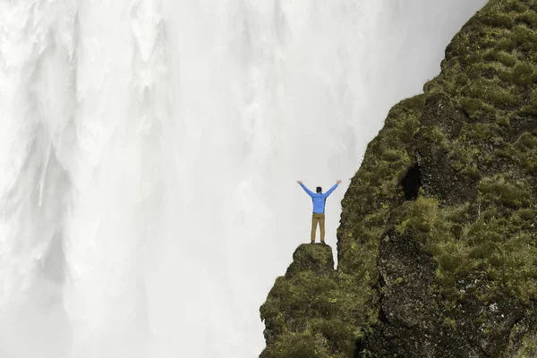 Junger Mann in blauer Jacke steht auf einer Klippe mit skogafoss Wasserfall im Hintergrund, Island. — Stockfoto