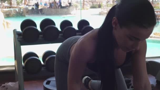 Beautiful girl with dumbbells. Girl shakes her hands in the gym. Sporty girl on the background of the pool and palms — Stock Video