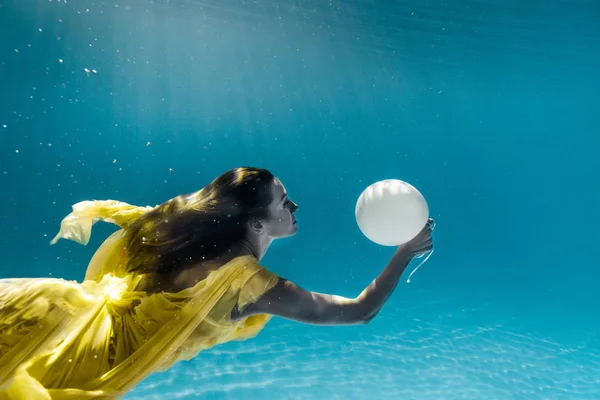 Underwater picture of beautiful young woman in dress with balloon swimming in swimming pool — Stock Photo