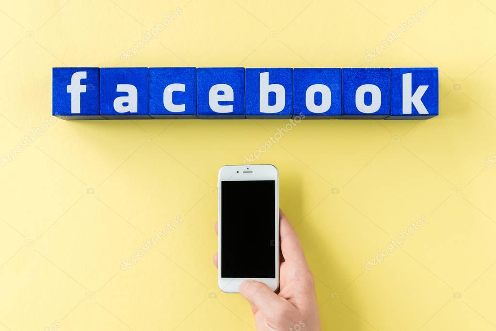 Facebook logo made from blue cubes and human hand holding smartphone on yellow surface