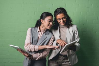 smiling african american and asian businesswomen working with digital tablets in front of green wall
