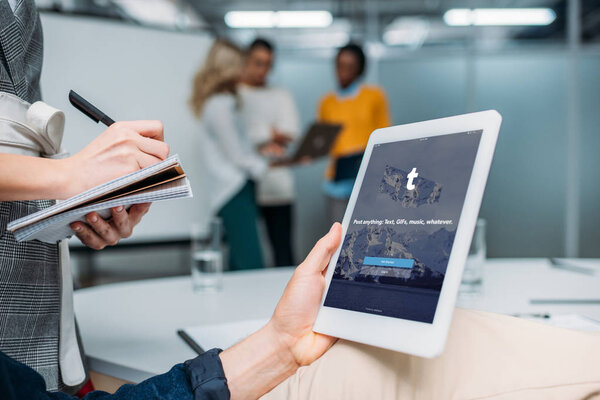 businessman holding tablet with tumblr on screen at modern office while colleague making notes