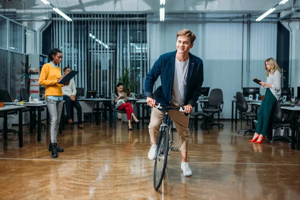 快乐的年轻商人骑自行车在现代办公室 而他的同事工作 — 图库照片