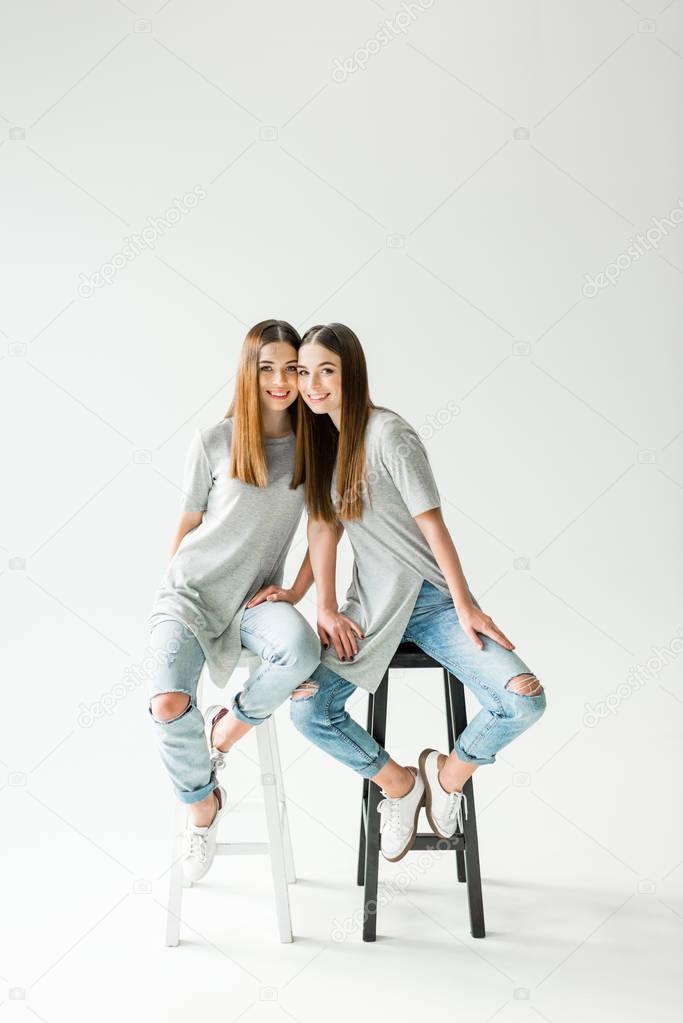 young cheerful twins in similar clothing looking at camera while sitting on chairs