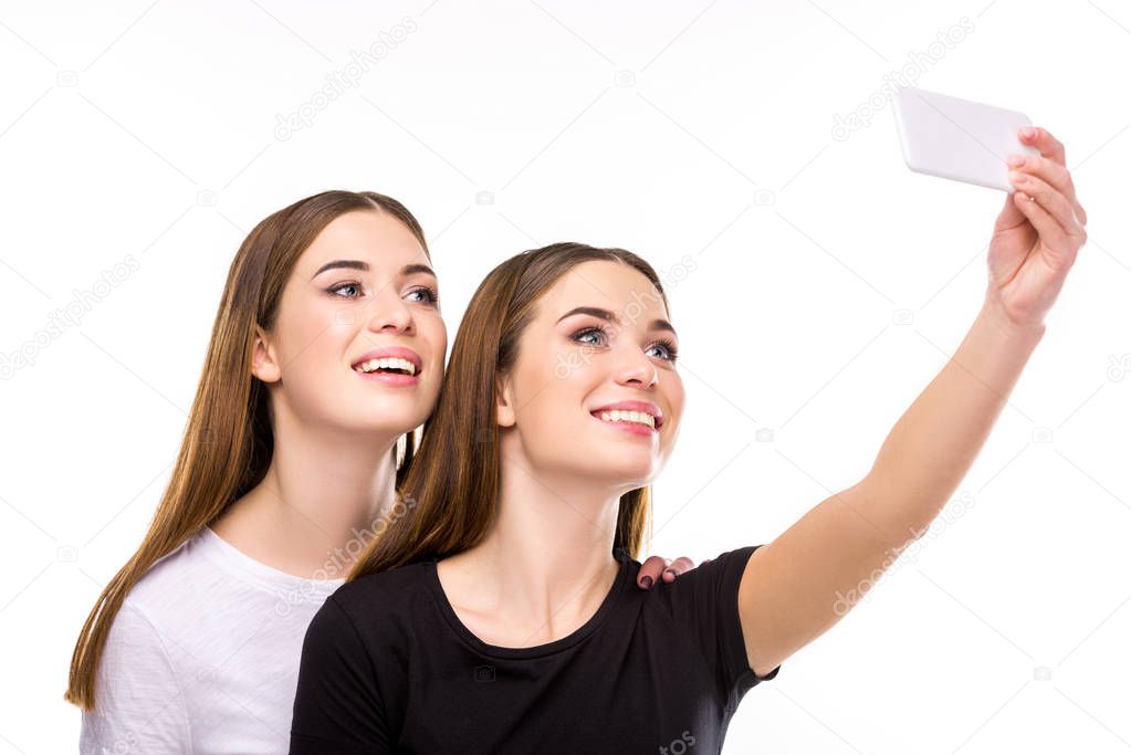 portrait of smiling twins taking selfie on smartphone together isolated on white