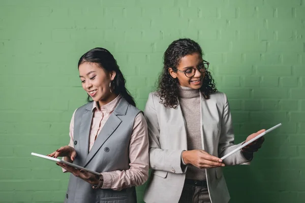 African american and asian businesswomen using digital tablets in front of green wall — Stock Photo