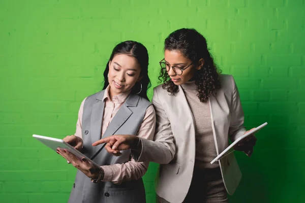 Mujeres de negocios afroamericanas y asiáticas que trabajan con tabletas digitales frente a la pared verde - foto de stock
