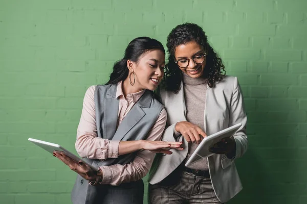 Souriantes femmes d'affaires afro-américaines et asiatiques travaillant avec des tablettes numériques devant le mur vert — Photo de stock