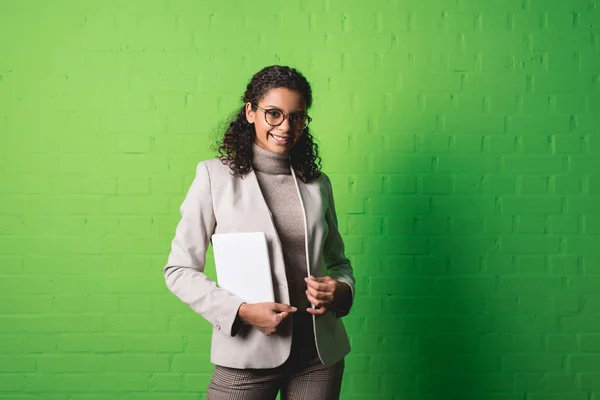 Joven mujer de negocios afroamericana sonriente con tableta digital frente a la pared verde - foto de stock