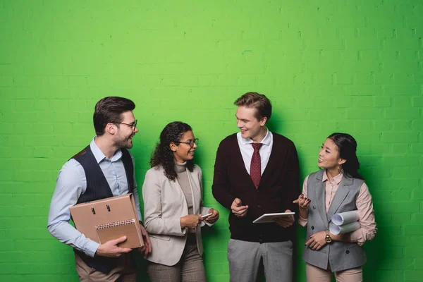 Young multiethnic businesspeople with digital devices and documents standing in front of green wall — Stock Photo