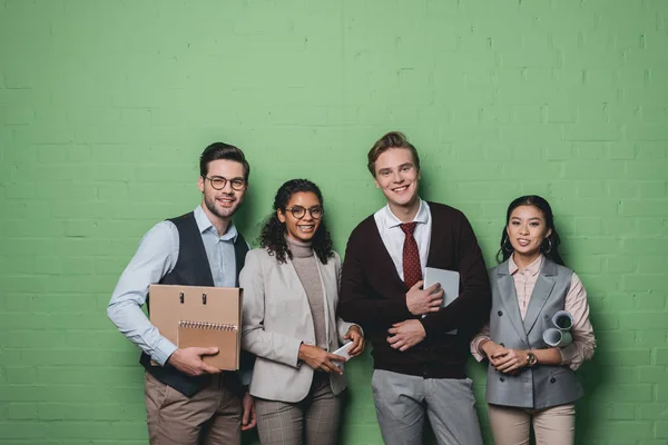 Young multiethnic businesspeople with digital devices and documents standing in front of green wall — Stock Photo