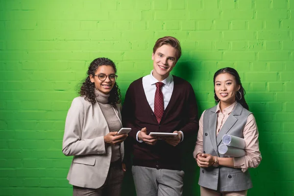 Joven hombre de negocios multiétnico y mujeres de negocios con gadgets y planos en frente de la pared verde - foto de stock