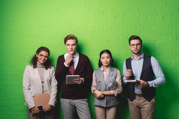 Jovens empresários multiétnicos com dispositivos digitais e documentos em pé em frente ao muro verde — Fotografia de Stock