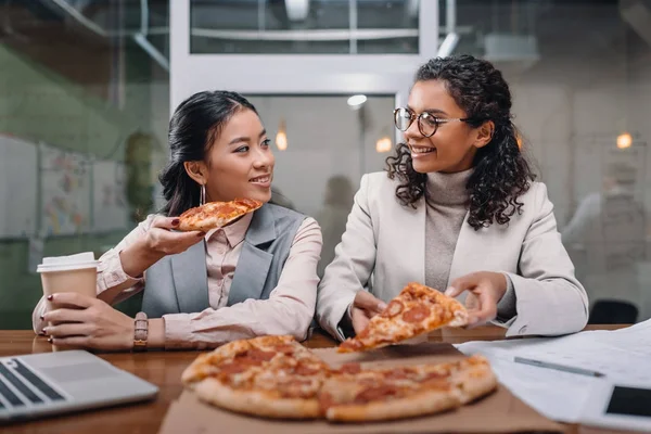 Мультиэтнические деловые женщины обедают в офисе, едят пиццу — стоковое фото
