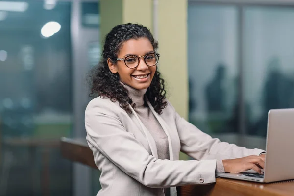 Молода усміхнена афроамериканська бізнес-леді в окулярах, що працюють з ноутбуком — стокове фото