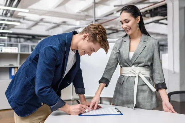 Handsome young man signing contract while businesswoman pointing at it — Stock Photo