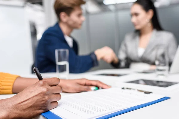 Recortado disparo de mujer afroamericana firma contrato mientras borrosa socios de negocios estrechando la mano en el fondo - foto de stock