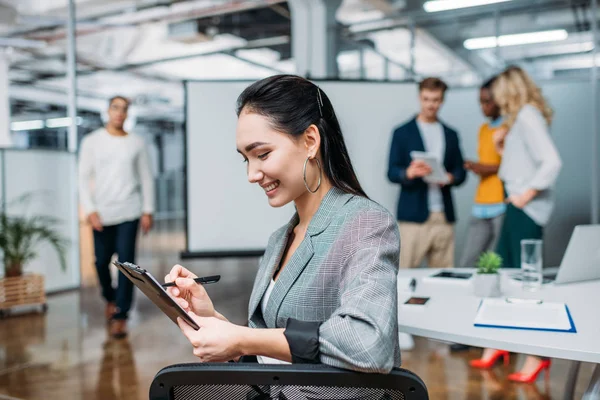 Young beautiful businesswoman writing on clipboard at modern office with blurred colleagues on background — Stock Photo