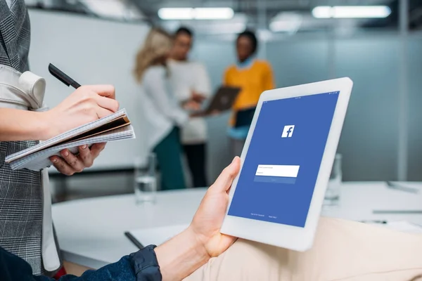 Hombre de negocios sosteniendo la tableta con facebook en la pantalla en la oficina moderna mientras colega toma notas - foto de stock