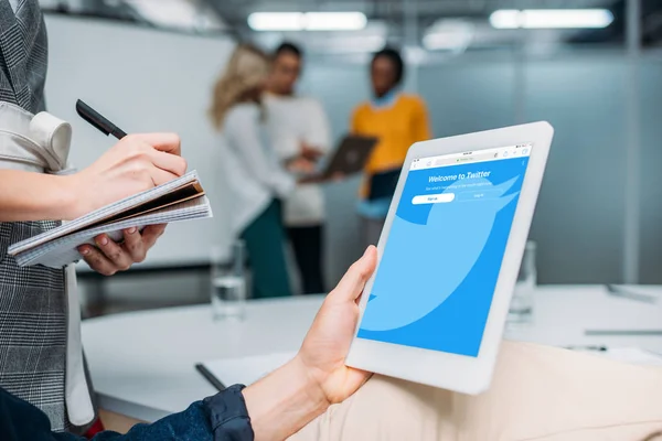 Hombre de negocios celebración de la tableta con twitter en la pantalla en la oficina moderna, mientras que el colega toma notas - foto de stock