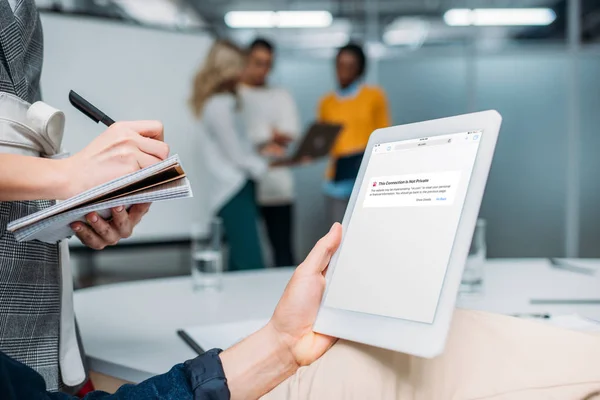 Hombre de negocios celebración de la tableta con la aplicación del navegador en la pantalla en la oficina moderna, mientras que el colega toma notas - foto de stock