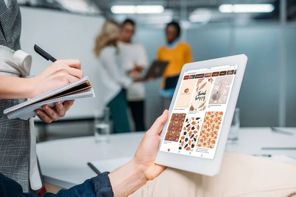 Hombre de negocios sosteniendo la tableta con pinterest en la pantalla en la oficina moderna mientras colega toma notas - foto de stock