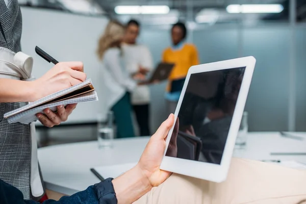 Hombre de negocios sosteniendo la tableta con pantalla en blanco en la oficina moderna mientras colega toma notas - foto de stock