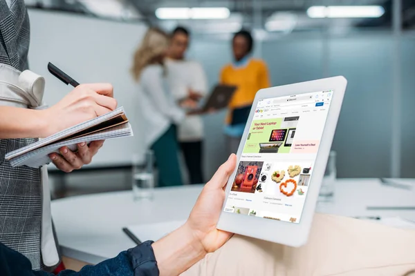 Hombre de negocios celebración de la tableta con ebay en la pantalla en la oficina moderna, mientras que el colega toma notas - foto de stock