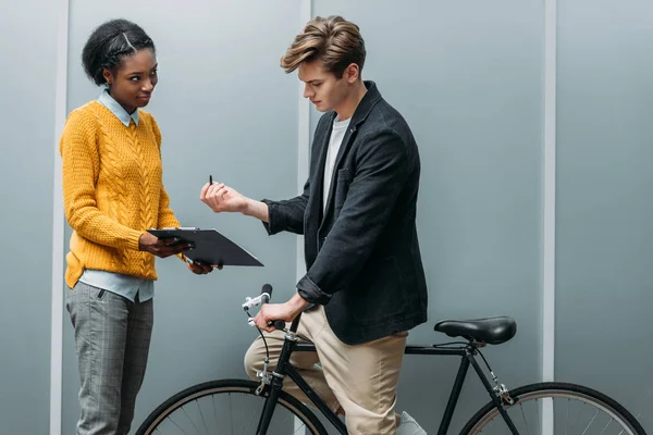 Handsome young businessman signing contract in hands of young african american colleague while sitting on bike — Stock Photo